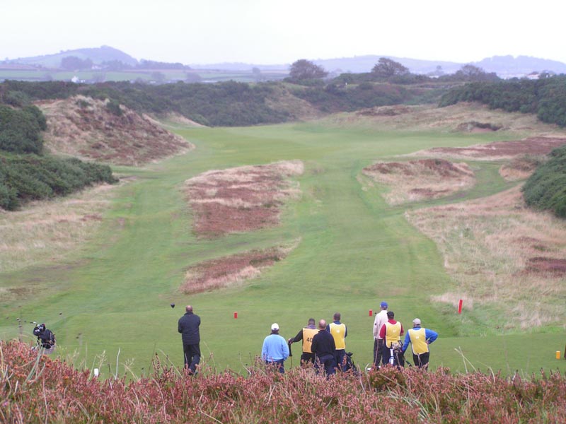 A view from the seventh tee shows the thirteenth fairway running through a valley between hills covered in weather and gorse. Can the golfer on the tee go long left with his tee ball to get a clear view of the green? Or is he best to lay-up near the base of the dune on the right...
